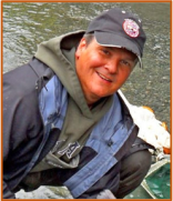 Headshot of Keith Parker in a jacket over a hoodie and wearing a dark baseball cap, crouching over gear with a shallow river in the background.