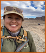 Maribel Lopez wearing a tan baseball cap and standing in a desert under a partly cloudy sky
