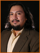 Headshot of Dr. Noel Novelo with long hair and beard, wearing a brown suit jacket in front of a black background.