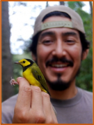 Xeronimo Castañeda holding a warbler. The bird is in focus, while Xeronimo, who is wearing a backward hat, and the trees behind him are out of focus.